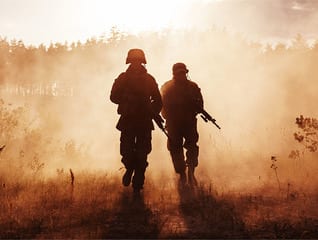 Two soldiers walking through a field with smoke coming from the ground.